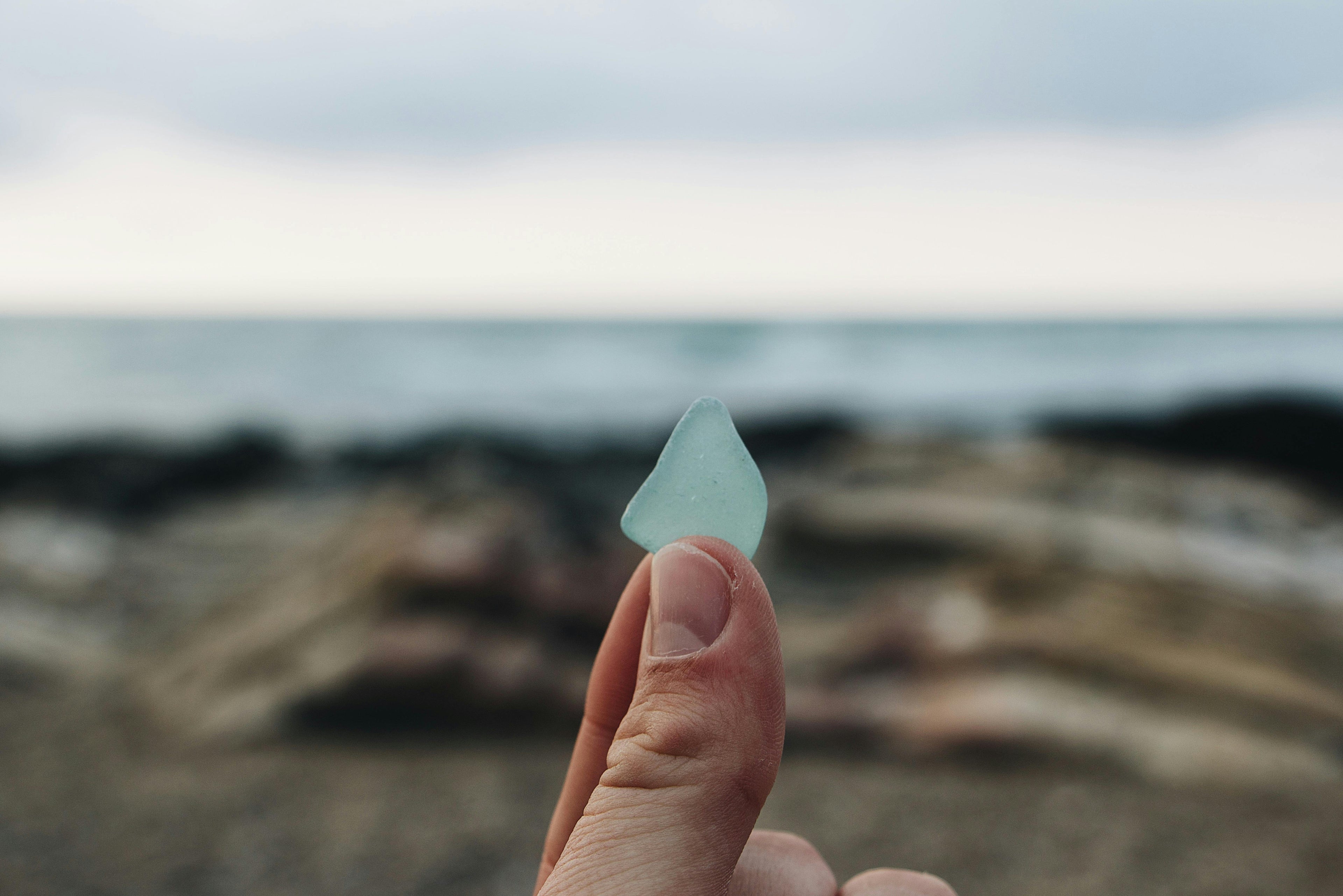 a picture of someone holding a teal piece of sea glass in their fingers in front of the ocean. 